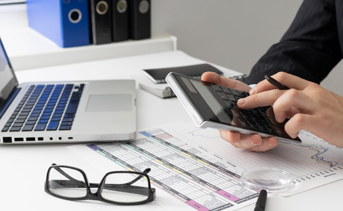 Person using a calculator and pen while analyzing financial documents on a desk with a laptop and glasses, illustrating financial consultancy and business solutions.