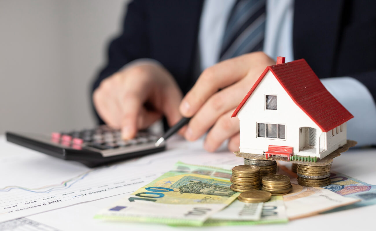 Man calculating finances with a calculator, surrounded by a model house and euro banknotes, representing real estate financing and business solutions.