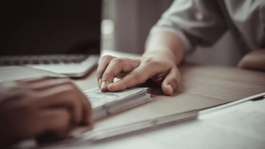 Hands exchanging cash on a table with a laptop and documents, illustrating financial transactions related to merchant cash advances and business funding.