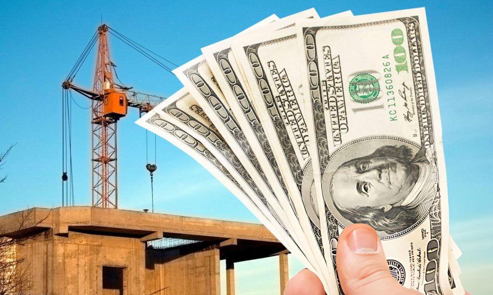 Construction loan lenders near me—hand holding cash in front of crane and concrete building site.